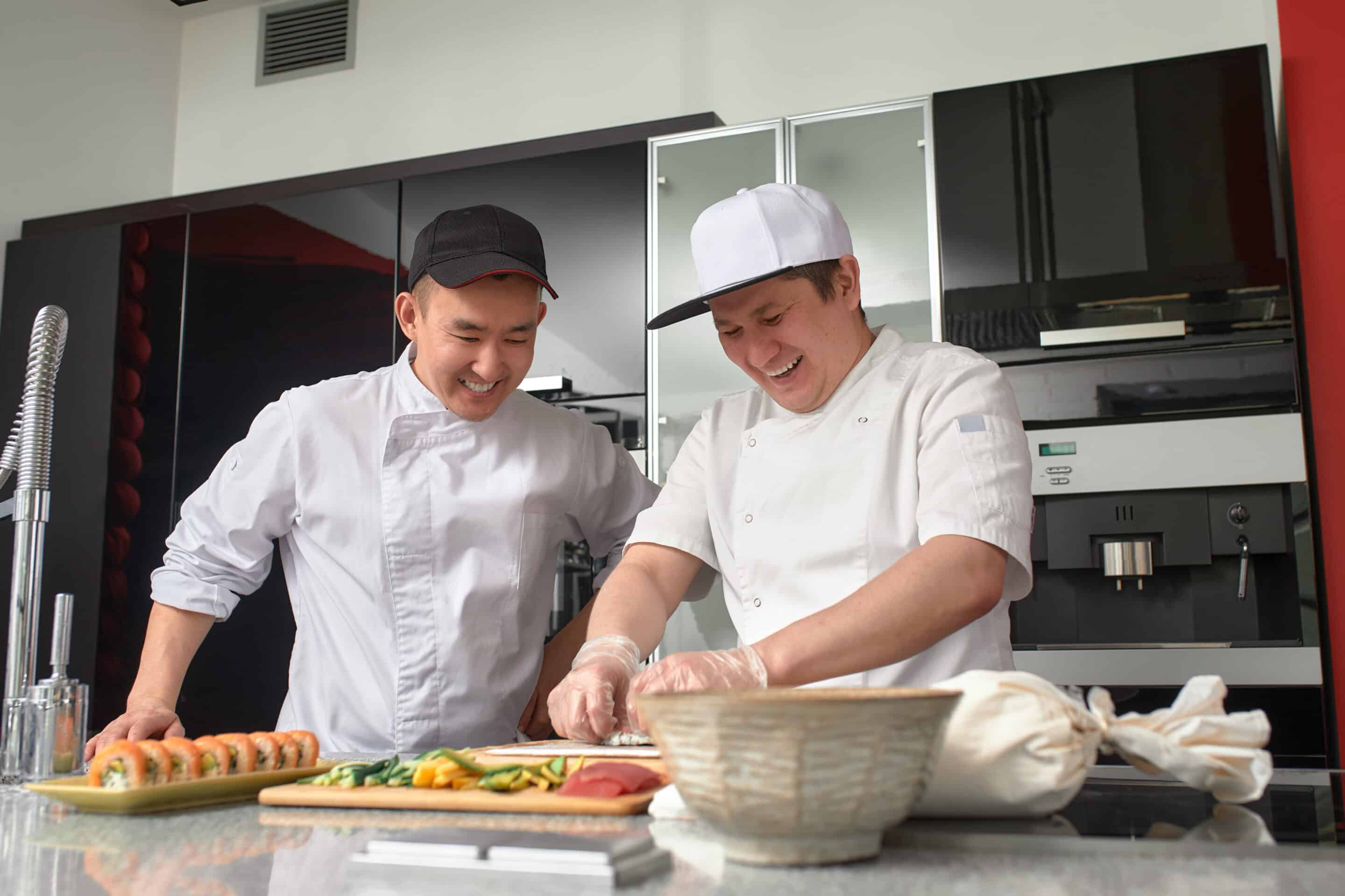 Two happy young asian chefs dressed in white uniform preparing traditional japanese sushi set in interior of modern professional kitchen.